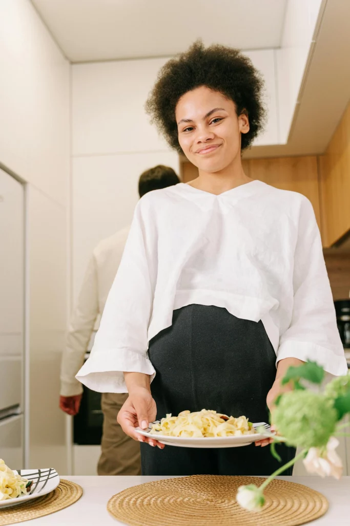 Schwangere Frau lächelt mit einem Teller Pasta in der Hand. Sie legt viel Wert auf gesunde Ernährung.