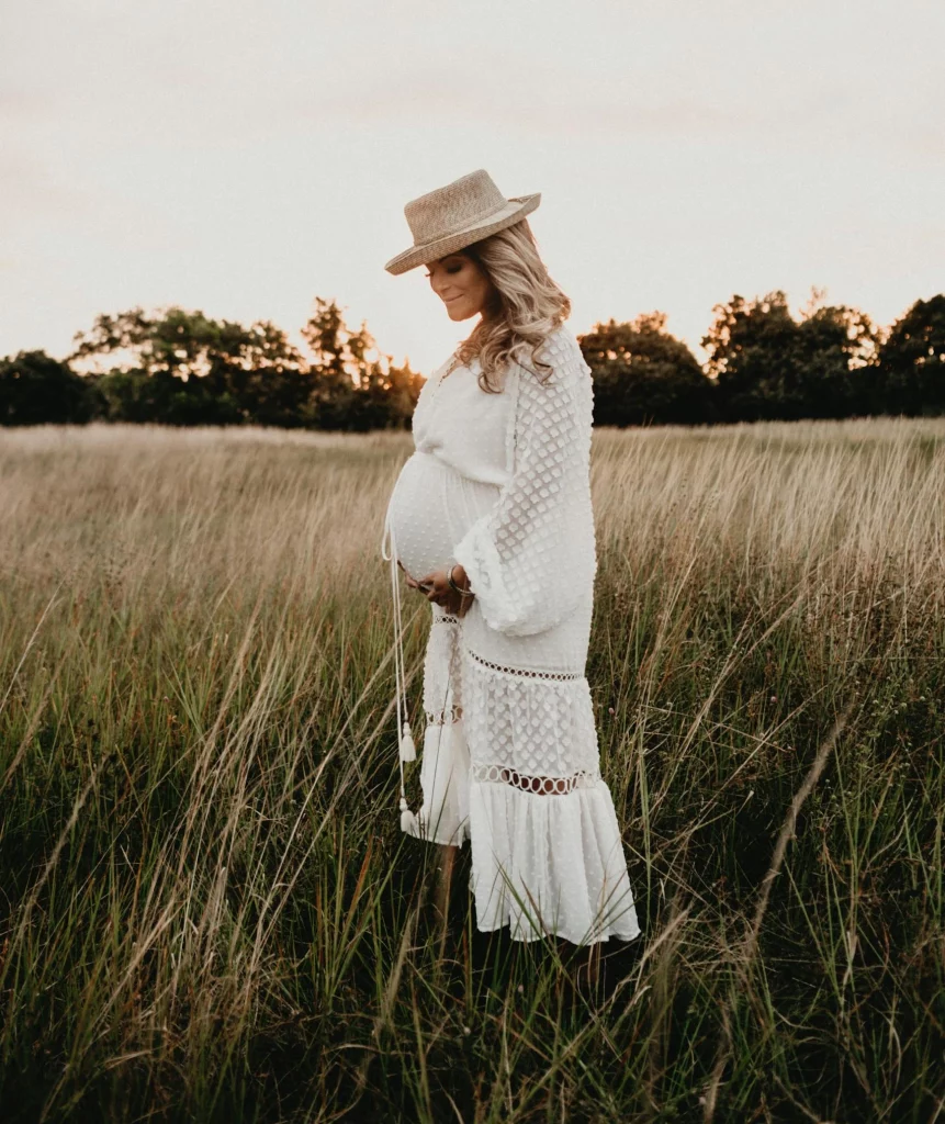 Frau steht in einem schicken weißen Kleid und Hut während ihrer Schwangerschaft auf einem Feld
